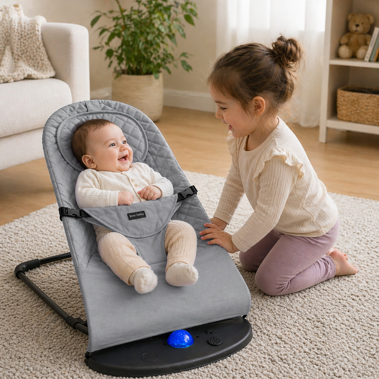 A baby sits in a gray new born baby bouncer chair on a beige rug, while a young girl kneels beside the chair in a bright living room. The bouncer has a black base with a small blue ball and a visible Baby Jug brand label.
